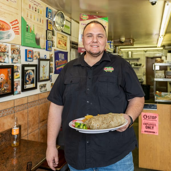 A man in a Little Mexico Chiquito shirt holds a large burrito with chips and vegetables.