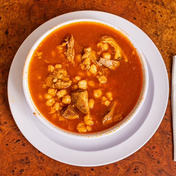 A bowl of Little Mexico Chiquito's pozole with red broth, meat, and hominy on a wooden table.