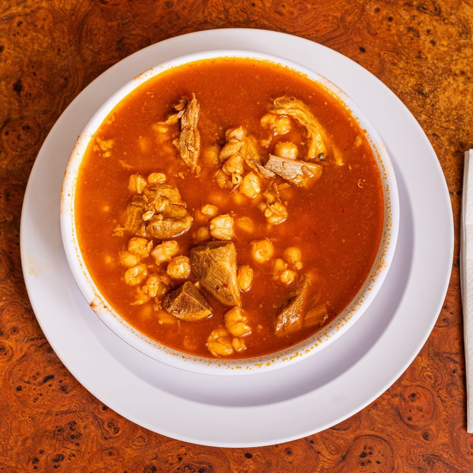 A bowl of Little Mexico Chiquito's pozole with red broth, meat, and hominy on a wooden table.