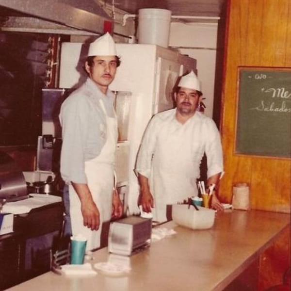 Two men in white chef hats and aprons stand behind a counter in a vintage Little Mexico Chiquito kitchen.