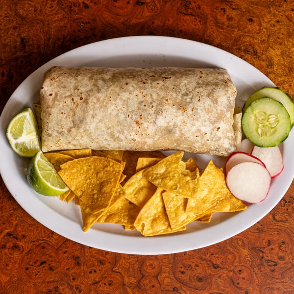 A large burrito with tortilla chips, lime wedges, cucumber slices, and radish slices on a white plate.