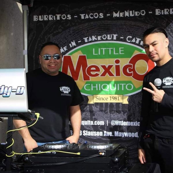 Two men in Little Mexico Chiquito shirts stand by a DJ setup and restaurant banner.