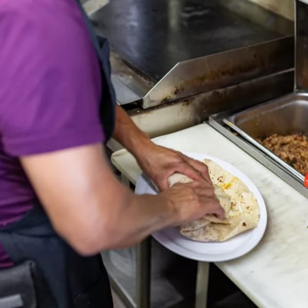 Chef's hands preparing a Mexican burrito on a white plate next to a large metal food warmer.