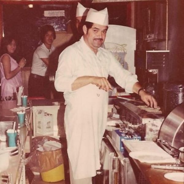 A chef in a white uniform and hat prepares food behind a counter at Little Mexico Chiquito.