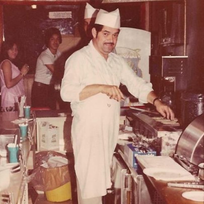 A chef in a white uniform and hat prepares food behind a counter at Little Mexico Chiquito.