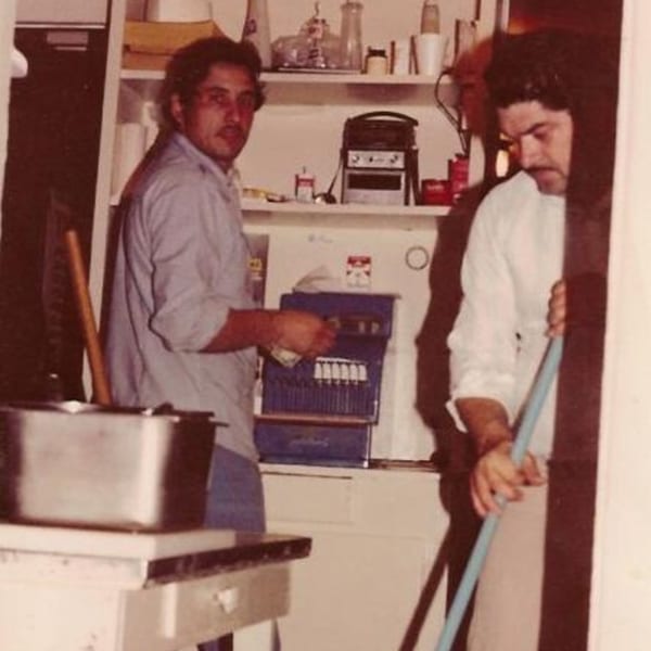 Two men in a kitchen, one holding money at a register, the other sweeping.