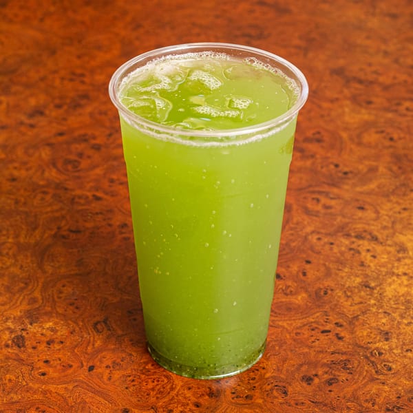 A vibrant green Mexican drink with ice and bubbles in a clear plastic cup on a wooden table.