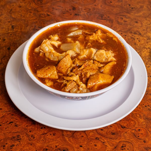 A bowl of menudo soup with tripe and hominy in a rich red broth on a wooden table.