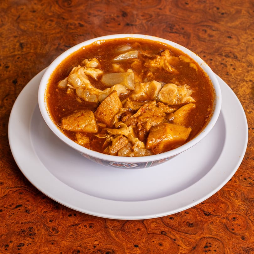 A bowl of menudo soup with tripe and hominy in a rich red broth on a wooden table.